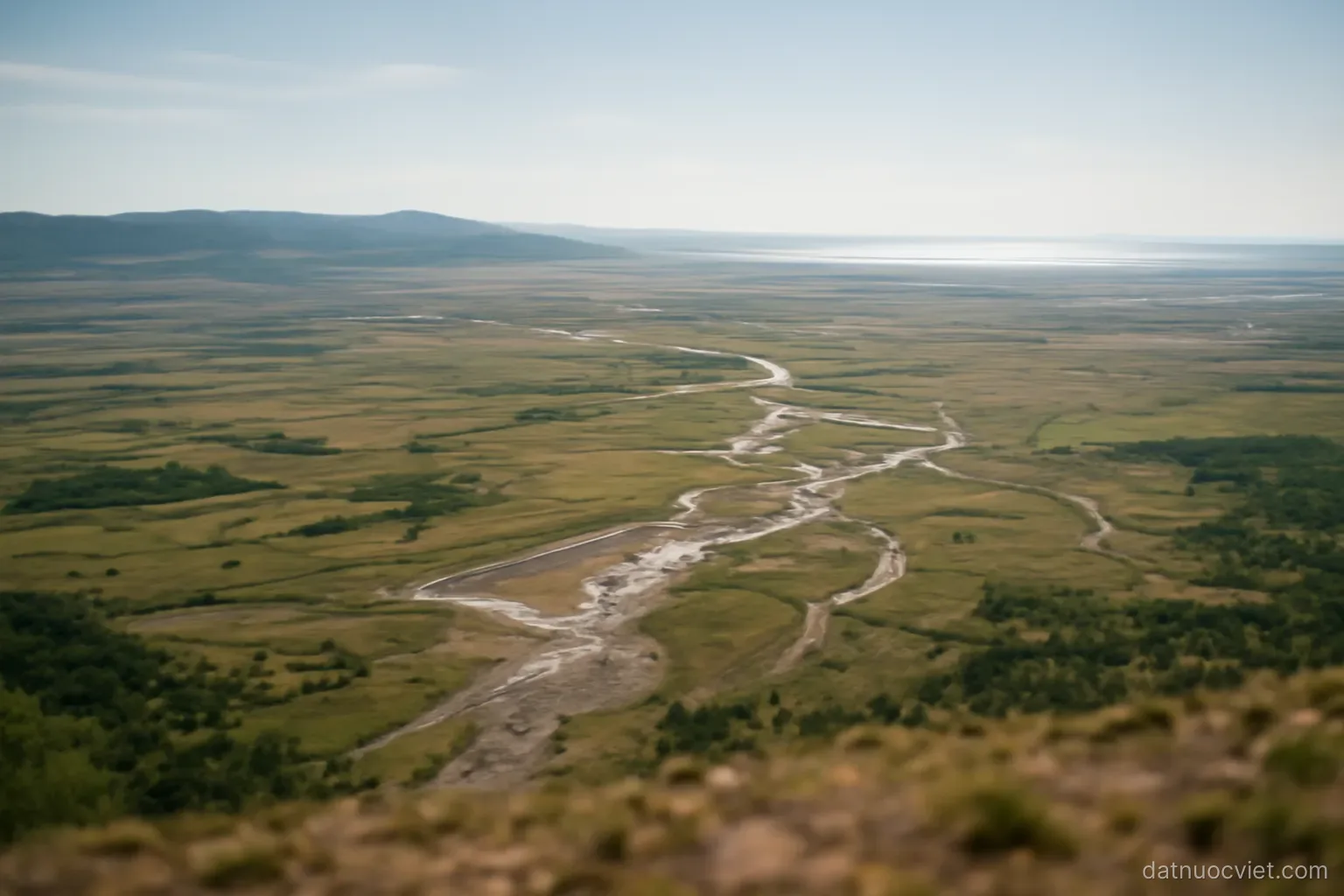 Broad alluvial plain seen from height with low hills still visible along one horizon and water glinting along the other