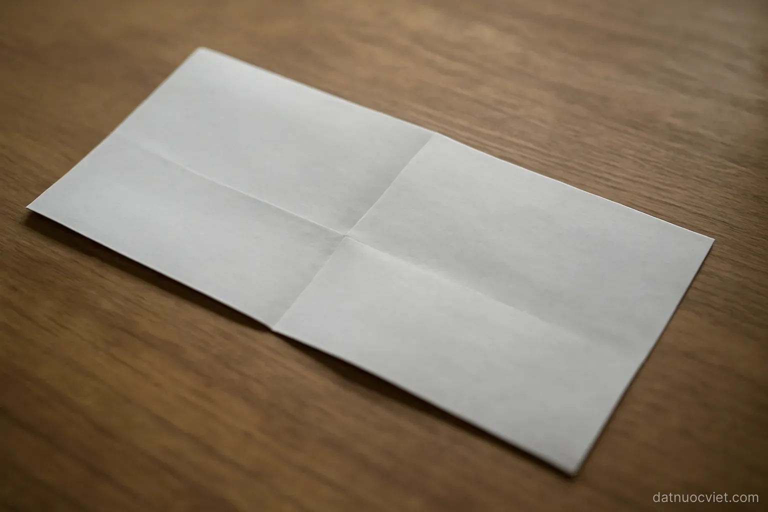 Long narrow sheet of paper on a table with one sharp crosswise crease and one faint lengthwise crease, both visible in raking light