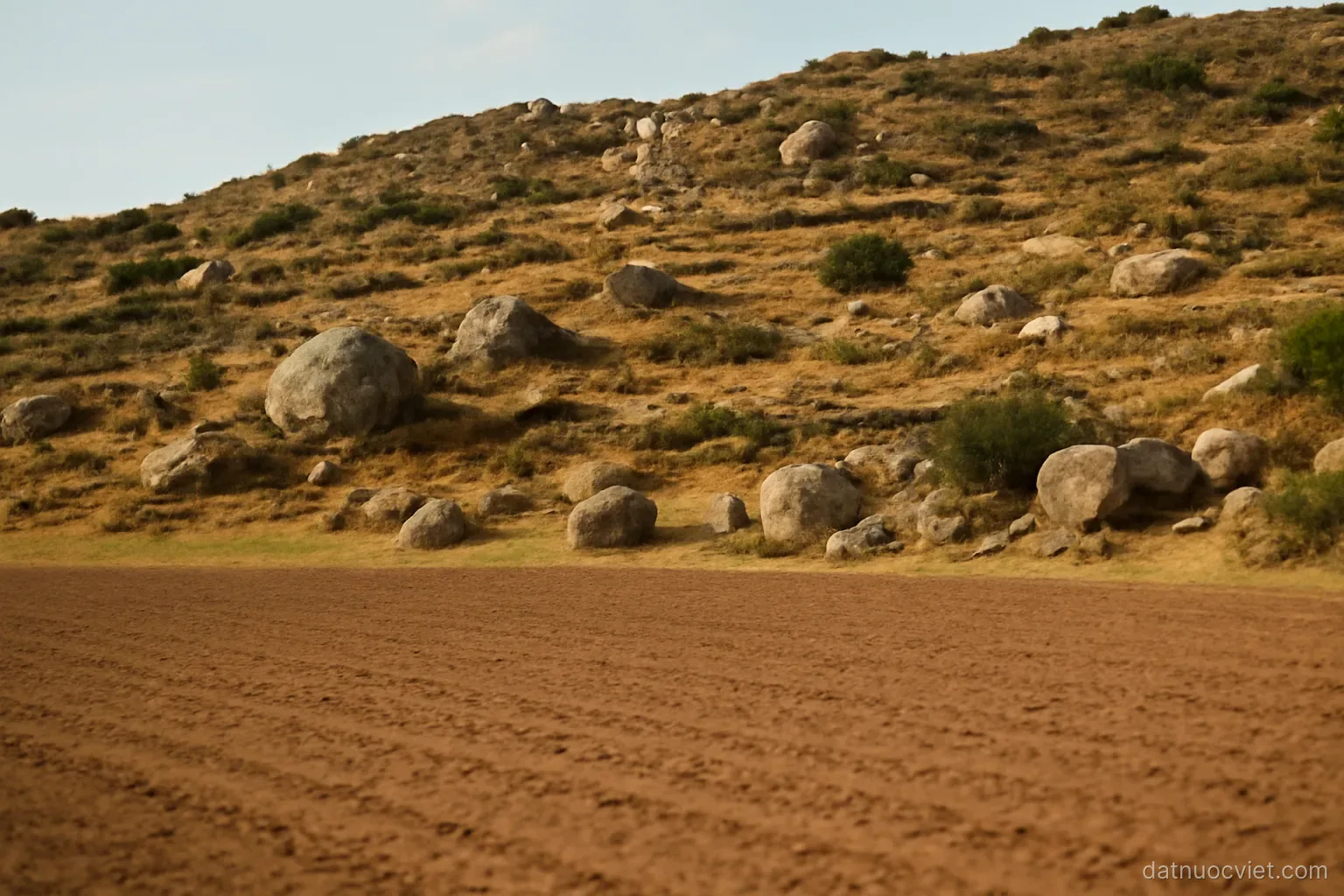 Gentle transition from flat cultivated ground into rising broken slopes, scattered boulders and terracing marking the change