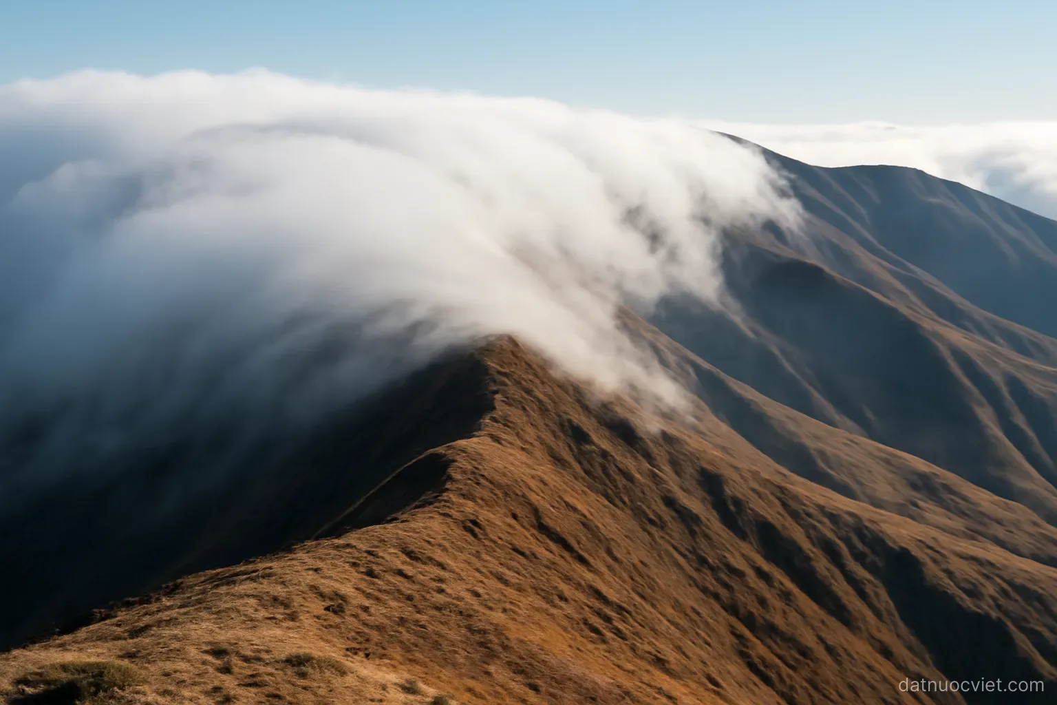 Cloud bank halted along a ridge crest with thin wisps spilling over the saddle, dry slopes visible on the far side